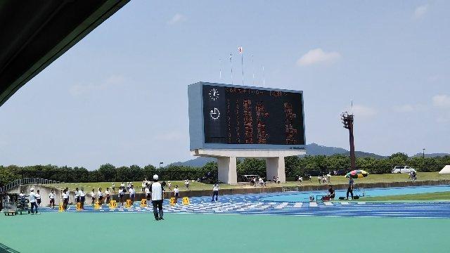 陸上競技場トラックのスタート位置でレーン毎に整列した児童達と周囲で見守る複数の指導者の様子と電光掲示板の写真