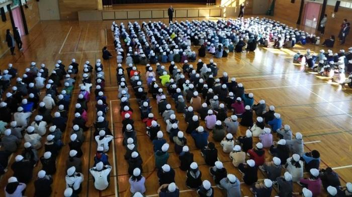 体育館内で帽子を着用し、綺麗に整列して座っている児童たちの写真