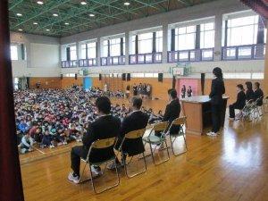 体育館のステージに横に並んで着席している6名の先生と演壇後ろに立つ先生と床に整列し座っている多くの児童達の様子の写真
