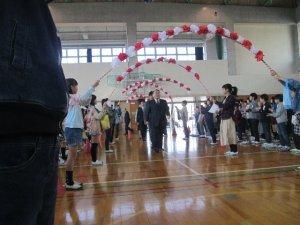 体育館で児童達が作った花のアーチの下をくぐり入場してくる先生たちの様子の写真
