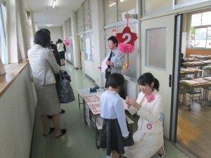 廊下で先生に胸に花をつけてもらっている新入生の児童と保護者の様子の写真