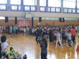 体育館で踊る先生、児童達と見守る左手前の児童達の様子の写真