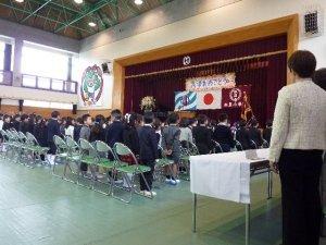 体育館のステージに「入学おめでとう」の横断幕と校旗国旗が飾られており並んだ椅子の前で起立している新入生の児童達の様子の写真