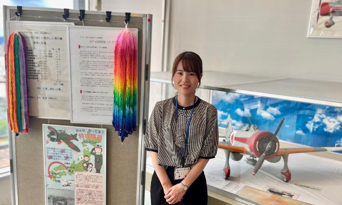 新館10階ロビーにて97式戦闘機と千羽鶴の前で説明している総務部総務課の薬谷さんの写真