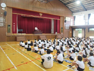 学校長が体育館の壇上で、あいさつについてほめている。