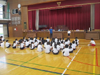 体育館で、交流する児童と4年生児童たちが集まって、話を聞いている。