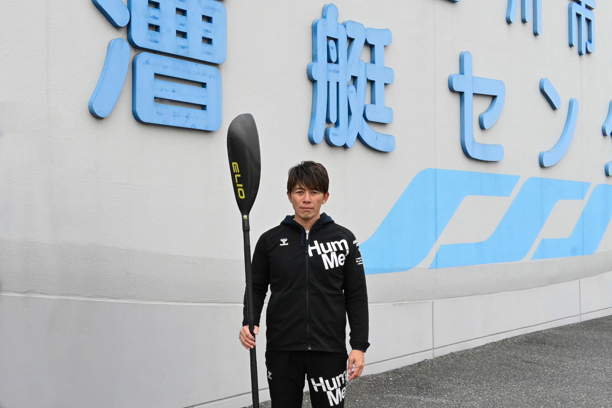 漕艇センターの看板前でパドルを持つ尾野藤さん