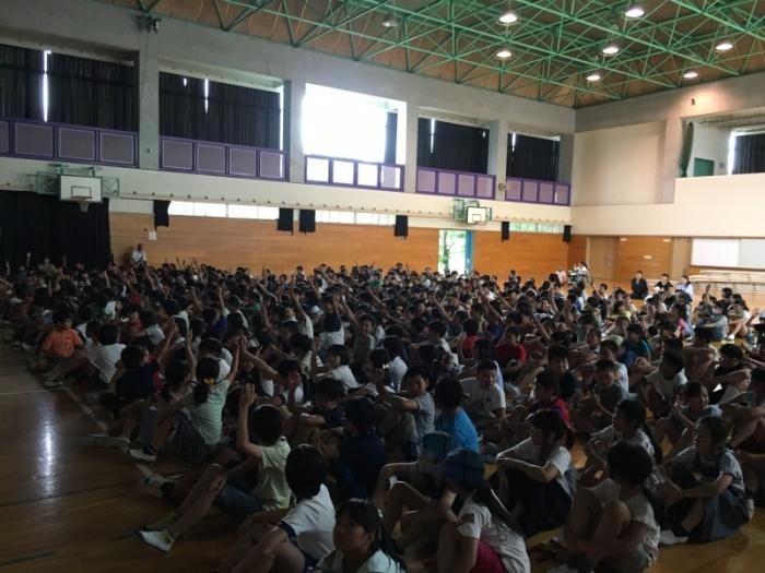 体育館に集まった4年生～6年生の児童達と保護者達が座って手を挙げている様子の写真