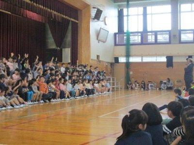体育館のステージ前に着席し手を挙げている1年生達と見守る上級生の児童達の写真
