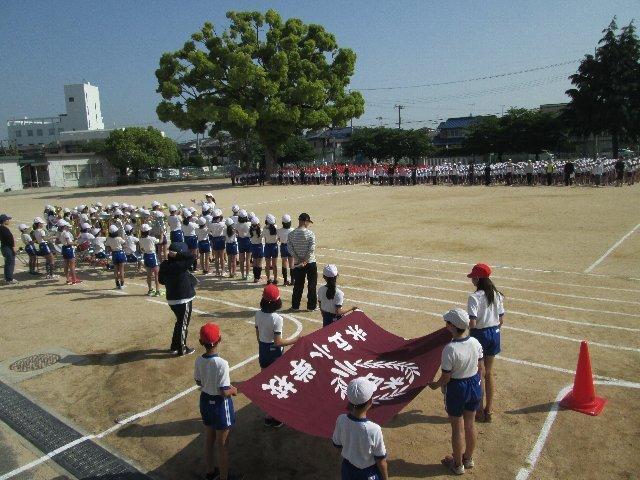 校庭で校旗を持つ6人の児童達と整列する全校児童と先生達の様子の写真