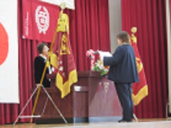 記念式典で、PTA会長から学校長に記念品目録を渡している。