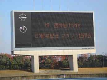 陸上競技場の電光掲示板に、「祝 西神吉小学校 120周年記念 マラソン記録会」と表示されている様子
