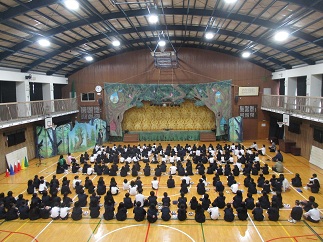 劇が始まる前の体育館。舞台装置を前に、児童が並んで座っている。