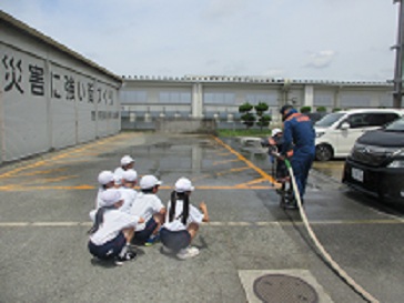 消防署で、耐火服を着た児童が、消防士に補助してもらいながらホースを使って放水している様子。