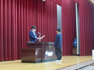 選任式の様子。代表の児童が体育館の壇上で校長先生から選任状を受け取っている。