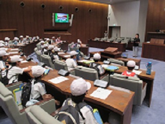 市役所の市議会議場で、児童たちが席に座らせてもらって、市議会についての話を聞いている。