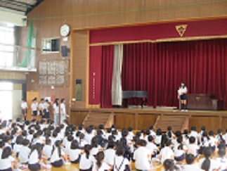 体育館で、委員会の代表が舞台に立ち、取り組みや目標を発表している。舞台横には、発表を待つ児童が並んでいる。