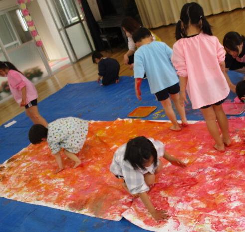 手に赤やオレンジの絵の具を付けた幼児が、大きな布中スタンプ遊びを楽しんでいるところ