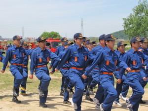 青い制服を着用し手を振りながら行進をする消防団員たちの写真