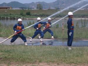 芝生の上の訓練で三人の消防団員が放水している写真