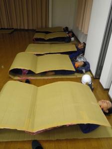 床の上に五人の消防団員が1列になりダンボール製の布団を敷いて眠りに就いている写真