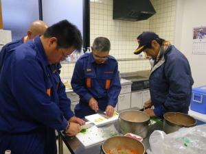 消防団員がキッチンのまな板の上で包丁を手に炊き出し訓練をしている写真