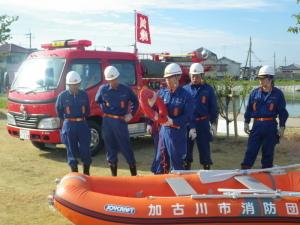 手前にボート、後ろに消防車が位置し、浮き輪を持った消防団員と周りの四人の消防団員が水難訓練をしている写真