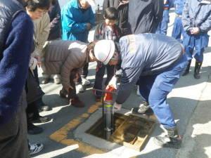 周囲に住民が集まり、中央で消防団員と住民がそれぞれ中腰になり消火栓使用法の指導をしている写真