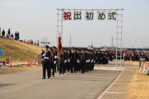 出初め式でプラカードや旗を持った先頭の団員に続いて行進をする消防団員たちの写真