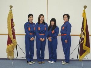 両サイドに旗が立ち中央で四人の女性消防団員が横一列に並んでいる写真