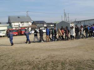 避難訓練で二人の消防団員の後に住民たちが続いて避難する写真